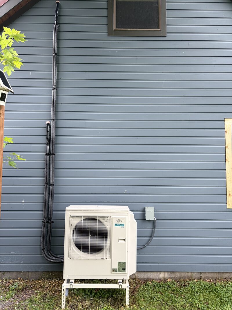 Outdoor mini-split unit installed on a stand beside a blue-sided West Michigan home